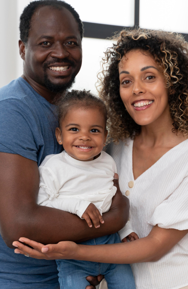 Uma família negra formada por pai, mãe e filha. Os três estão se abraçando, sorrindo para frente, enquanto a criança se encontra no colo do pai